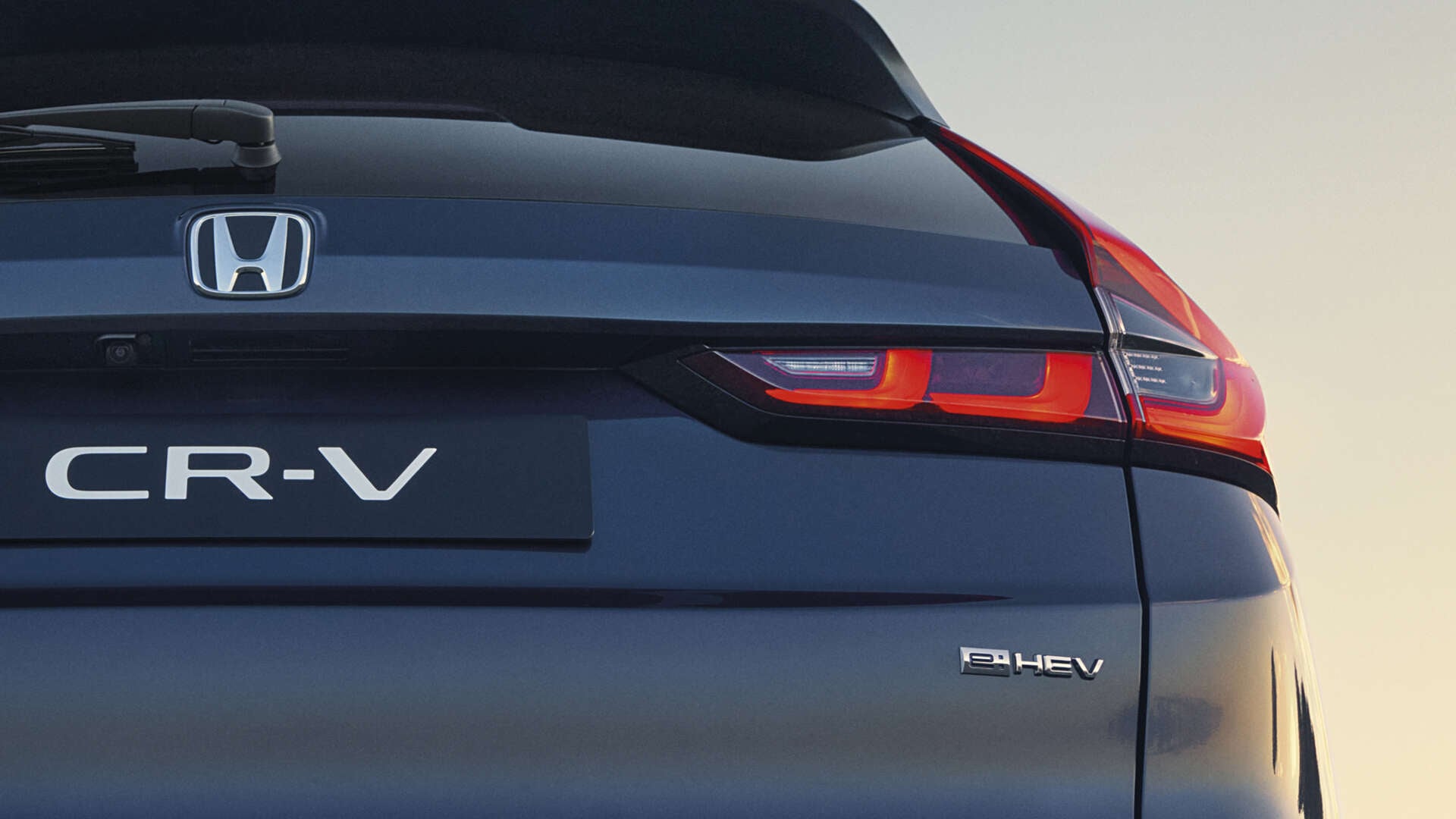 Detailný pohľad zozadu na tmavomodrú Honda CR-V e:HEV s logom Honda, nápisom CR-V a osvetlenými charakteristickými zadnými LED svetlami na pozadí oblohy pri západe slnka.