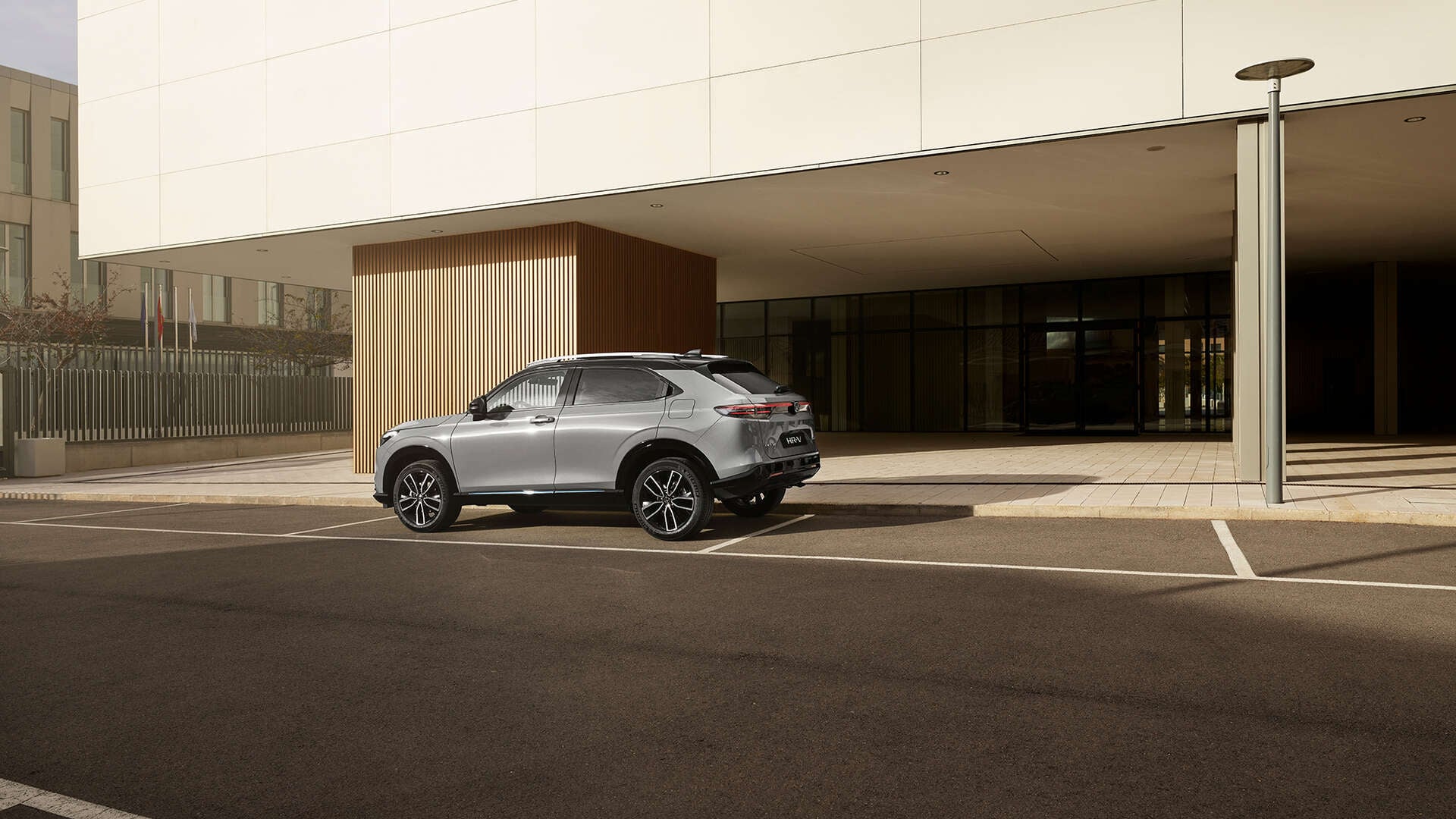 Strieborná Honda HR-V je zaparkovaná na vonkajšom parkovacom mieste pred modernou budovou so sklenenými stenami a zvislým dreveným obložením, fotografovaná zo zadného trojštvrťového uhla za denného svetla.